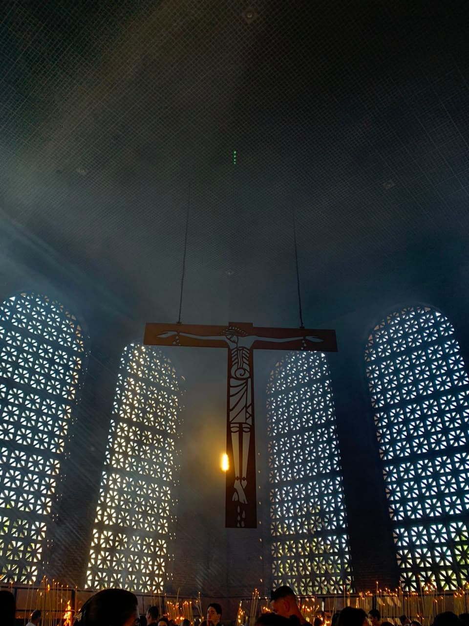 O grande crucifixo suspenso dentro da Basílica de Aparecida, entre janelas rendadas que deixam a luz entrar, com velas acesas lá embaixo