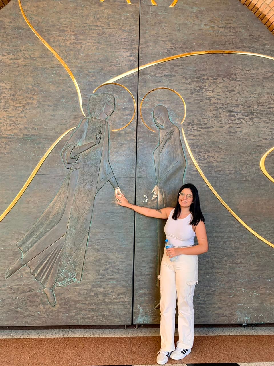 Taíssa encostando a mão nas grandes portas de bronze da Basílica de Aparecida, com anjos em relevo e traços dourados