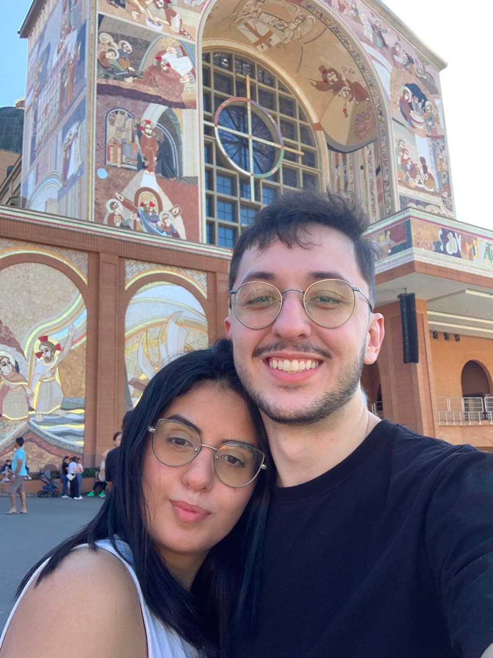 Selfie de Taíssa e Tiago sorrindo diante da fachada de mosaicos da Basílica de Aparecida
