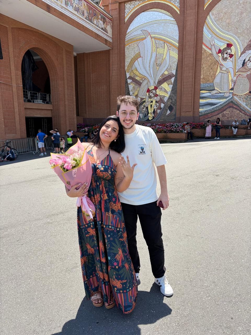Taíssa e Tiago sorrindo diante da fachada de mosaicos da Basílica de Aparecida, ela com um buquê de rosas