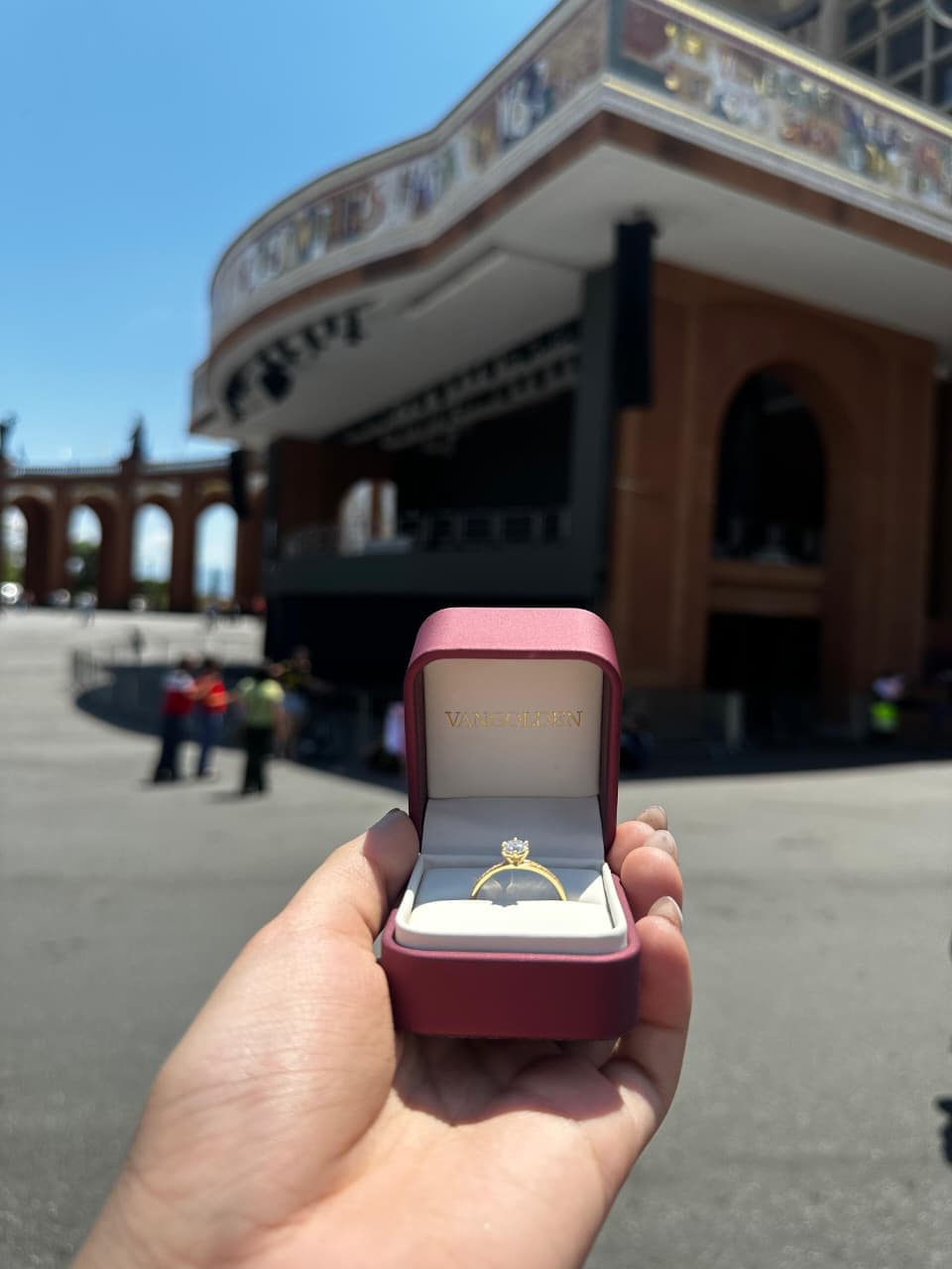 A mão do Tiago segurando a caixinha aberta com a aliança, diante dos arcos da Basílica de Aparecida